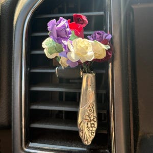 May include: A decorative car air vent accessory featuring a repurposed silver spoon as a vase. It holds a small bouquet of paper flowers in shades of purple, red, yellow, and green, adding a touch of colour to the car's interior.