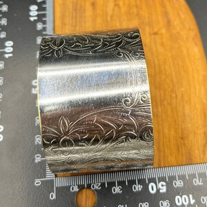 May include: Silver napkin ring with an ornate floral design. The ring is approximately 5 cm in diameter.