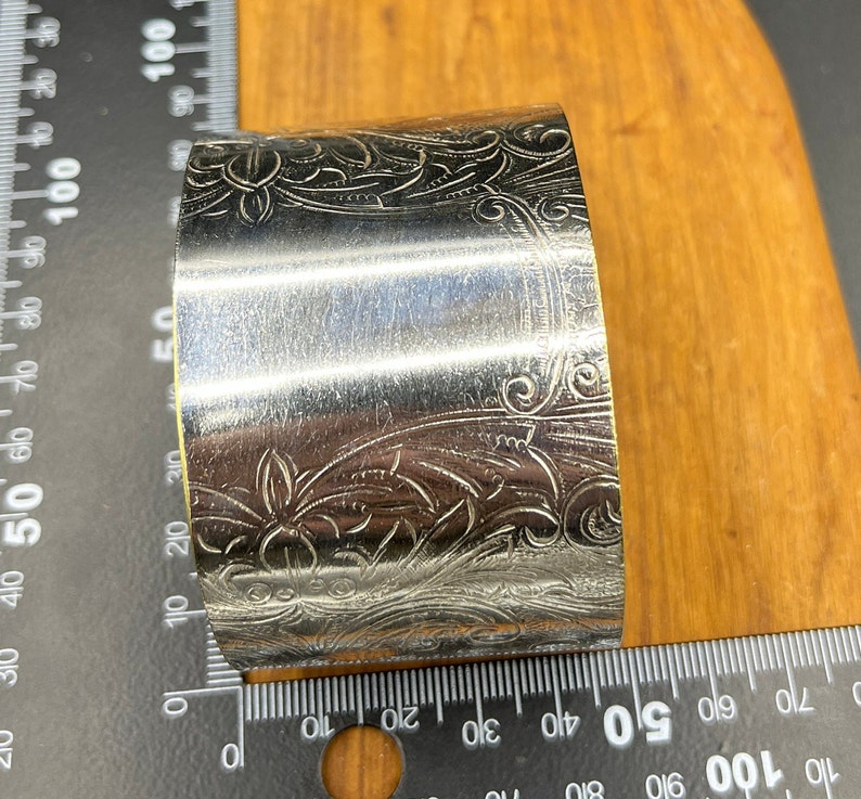 May include: Silver napkin ring with an ornate floral design. The ring is approximately 5 cm in diameter.