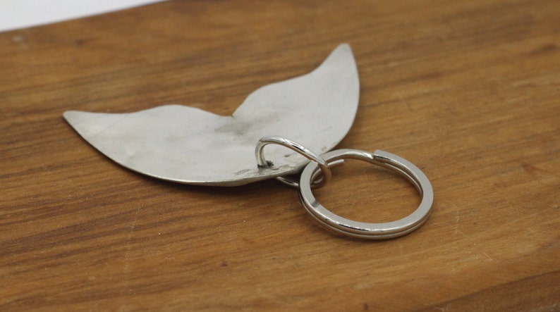 May include: Silver whale tail keychain with a silver ring. The whale tail is a simple design with a smooth surface.