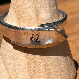 May include: A silver adjustable ring featuring the Leo zodiac symbol. The ring is displayed on a wooden stand. The band has a textured finish, and the Leo symbol is engraved. The adjustable design allows for a comfortable fit, suitable for various finger sizes.