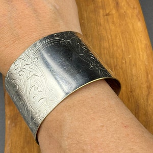 May include: A silver cuff bracelet featuring an intricate floral design. The bracelet is wide and has a smooth, polished finish.
