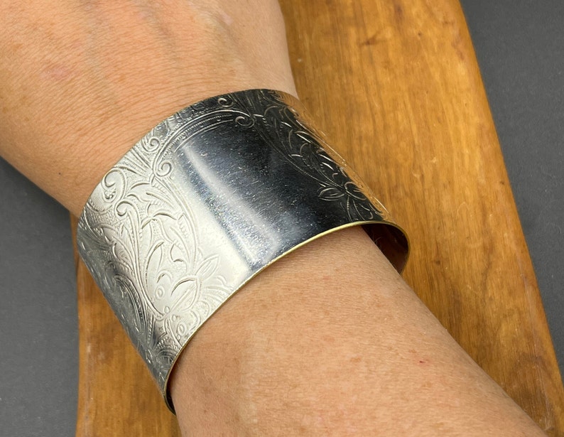 May include: A silver cuff bracelet featuring an intricate floral design. The bracelet is wide and has a smooth, polished finish.