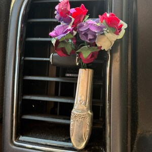 May include: A silver spoon repurposed as a car air vent vase, holding a bouquet of colourful artificial flowers. The flowers include red, purple, and white blooms with green leaves. The spoon has ornate detailing.