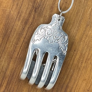 Puede incluir: Un collar de horquilla de plata con un diseño decorativo en el mango. Los dientes de la horquilla están doblados para formar un corazón.