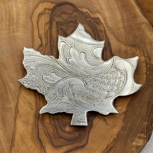 May include: A silver-colored maple leaf-shaped brooch with intricate engraved details. The brooch has a detailed pattern of swirling lines and textures, creating a vintage or antique aesthetic. The brooch is displayed on a wooden surface.