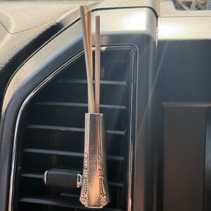 May include: A silver-coloured car air vent diffuser with a decorative floral design. The diffuser holds several light-coloured wooden reeds. The diffuser is placed in a black car air vent.