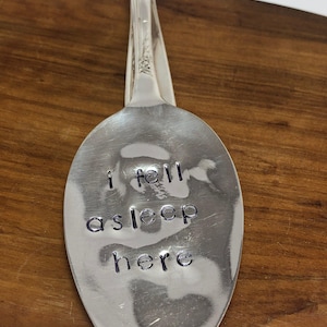 Pode incluir: Uma colher de prata com o texto estampado "i fell asleep here". A colher está sobre uma superfície de madeira. O cabo da colher está dobrado e tem um desenho floral.