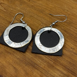May include: A pair of silver earrings with a black leather backing. The earrings are circular with a decorative design.