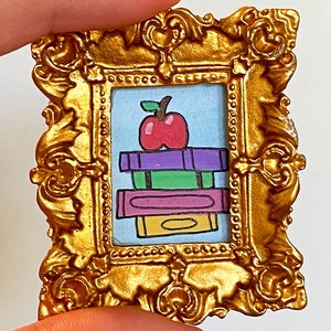 May include: An ornate gold frame containing a colourful drawing of a stack of books with an apple on top. The apple is red with a green stem and leaf. The books are purple, green, pink and yellow.