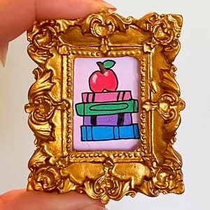 May include: An ornate gold frame with a pink background and a drawing of a stack of books with a red apple on top. The books are drawn in different colours, including green, blue, and purple.