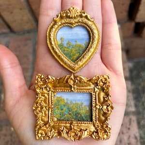 May include: Two miniature gold-colored frames, one heart-shaped and one rectangular, each containing a small landscape painting. The paintings depict a field of yellow flowers under a blue sky. The frames have ornate, detailed designs.