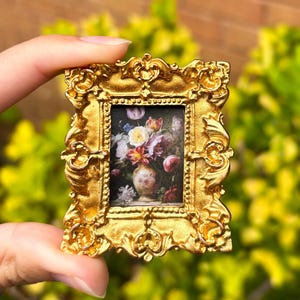 May include: A miniature gold-colored framed painting featuring a still life of flowers in a vase. The frame has an ornate, detailed design. The painting's colors include red, yellow, and white.