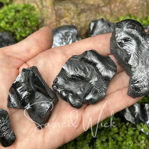 May include: Several pieces of raw, black obsidian are held in a hand, with a green, mossy background. The obsidian pieces have a shiny, reflective surface and irregular shapes. The text "Sacred Wick" is visible in the image.
