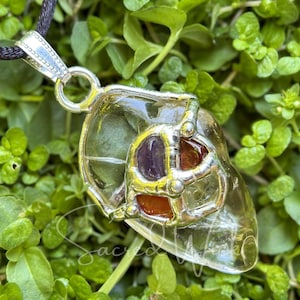 May include: A clear quartz crystal pendant with a silver cage design. The cage features three smaller crystals: amethyst, carnelian, and clear quartz. The pendant is on a black cord.