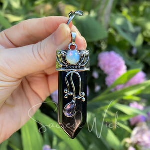 May include: A black pendant with a silver-colored top and accents, featuring a white opal-like stone and a dangling amethyst-colored crystal. The pendant is held against a backdrop of green foliage and pink flowers.