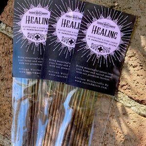 Könnte beinhalten: Drei Packungen Räucherstäbchen mit einem lila und schwarzen Etikett, auf dem "Healing Kaffir Lime & Dark Amber 10 Handcrafted Sticks" steht. Das Etikett enthält auch ein kleines Kreuz und ein Sonnenstrahlen-Design. Der Text "Bring meaningful intent into your home and sacred space with our premium incense. Artisan Witchcrafts By www.sacredmists.com" steht ebenfalls auf dem Etikett.