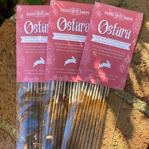 May include: Three packages of incense sticks labeled "Ostara Dragonfruit & Sandalwood 10 Handcrafted Sticks". The packages are pink with white floral designs and a white rabbit illustration. The text "Artisan Witchcrafts By www.sacredmists.com" is printed on the bottom of the packages.