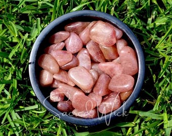 May include: A black bowl filled with polished, pink-orange gemstones. The bowl sits on a bed of green grass. The gemstones have a smooth, rounded shape and vary in size. The text "Sacred Wick" is written on the bottom right of the bowl.