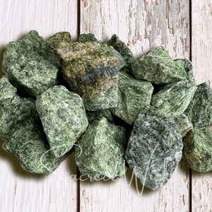 May include: A pile of rough, green serpentine stones. The stones vary in size and shape, with a mix of light and dark green hues. The background is a weathered, white-painted wooden surface.