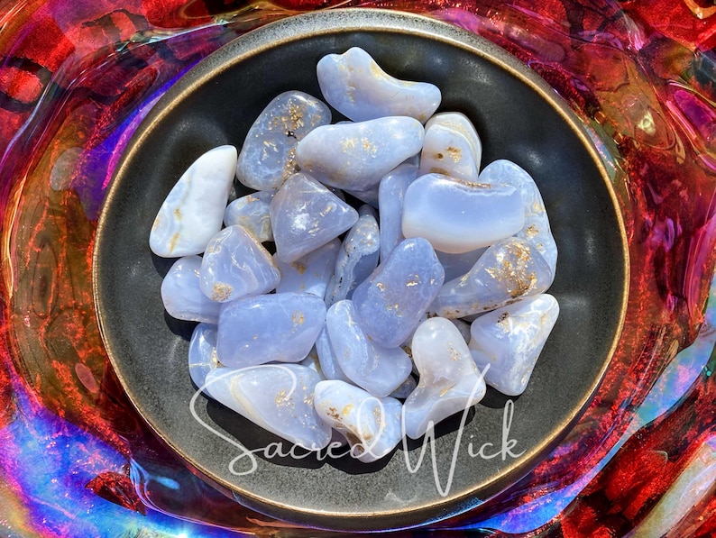 An assortment of tumbled blue chalcedony stones in a bronze bowl basking in sunlight, showcasing their soft, soothing blue tones and gentle energy known for promoting calm, emotional balance, clear communication, and spiritual connection.