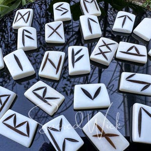 May include: A set of 24 white bone rune stones with black markings. The runes are arranged in a semi-circle on a black surface. The runes are used for divination and magic.