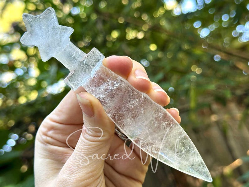 Clear quartz crystal athame with star handle held between fingers, showing clarity against greenery background.
