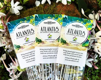 May include: Three packages of incense sticks with a beach scene and the text "ATLANTIS SEA GRAPES & SAND VERBENA" on the front. The text "SACRED MISTS SHOPPE" is also on the front of the packages. The packages are surrounded by white flowers.