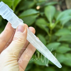Clear quartz crystal athame held between fingers, angled to display depth with greenery behind.