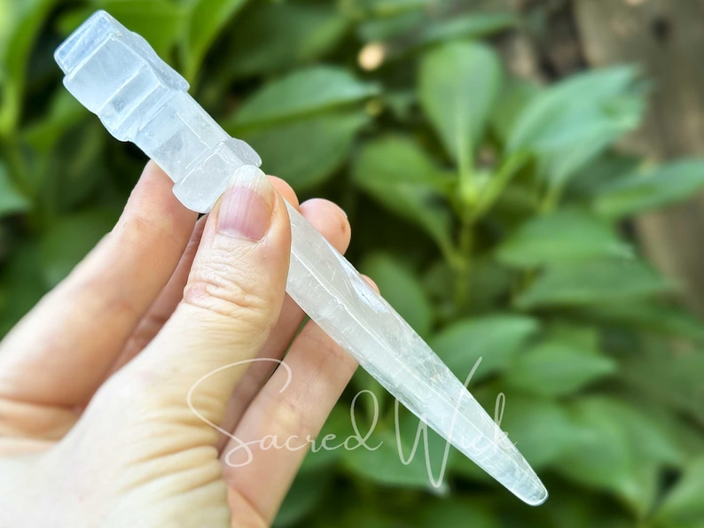 Clear quartz crystal athame held between fingers, angled to display depth with greenery behind.