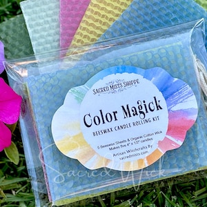 May include: A clear plastic bag contains a candle-making kit with colorful beeswax sheets and organic cotton wicks. The label reads "Color Magick" and "Beeswax Candle Rolling Kit". The kit makes five 4" x 1/2" candles. A pink and purple flower is in the background.