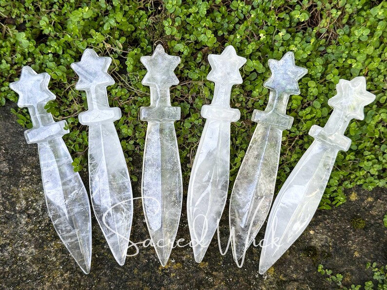 Six clear quartz crystal athames with star handles lying on greenery with a rock at the bottom.