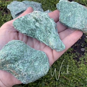May include: Green serpentine rough stone, a type of gemstone, in various sizes and shapes. The stones are in a hand, showing their natural texture and color.