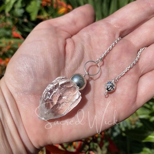 Könnte beinhalten: Ein klares Quarzkristall-Pendel mit einer silbernen Kette und einer silbernen Kugel an der Spitze. Das Pendel wird in einer Hand gehalten.