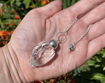 May include: A clear quartz crystal pendulum with a silver chain and a silver ball at the top. The pendulum is held in a hand.