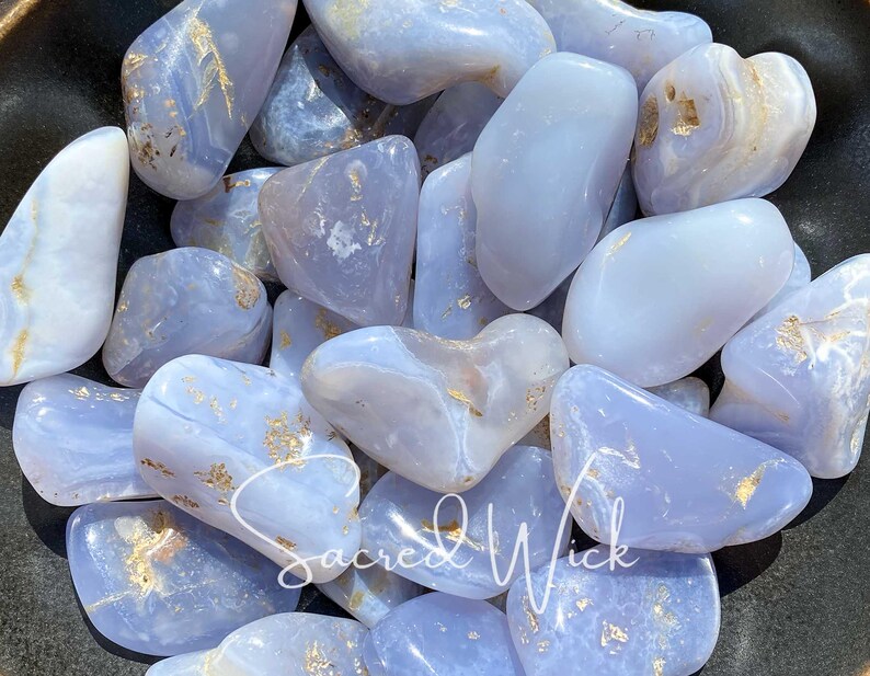 A close up of an assortment of tumbled blue chalcedony stones in a bronze bowl basking in sunlight, showcasing their soft, soothing blue tones and gentle energy known for promoting calm, emotional balance, clear communication, and spiritual connection.