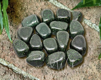 Puede incluir: Un grupo de piedras rodadas de aventurina verde pulida. Las piedras son lisas, redondeadas y salpicadas de inclusiones brillantes. Están dispuestas sobre una superficie marrón texturizada, con follaje verde en el fondo. El texto "Sacred Wick" es visible.
