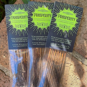 May include: Three packages of incense sticks labeled "Prosperity" with the text "Egyptian Kyhpi & Almond 10 Handcrafted Sticks" and a dollar sign. The incense sticks are brown and have a blue tint. The packages are on a brown surface.