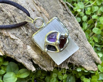 May include: A silver pendant with a selenite crystal and three gemstones: amethyst, garnet, and black onyx. The pendant is on a black cord and is resting on a piece of driftwood.