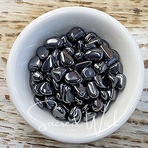 May include: A white ceramic bowl filled with polished, dark gray hematite stones. The stones have a smooth, reflective surface. The text "Sacred Wick" is written in a cursive font at the bottom of the bowl.