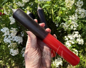 May include: Two black and red taper candles. The candles are held in a hand. The candles are in front of a background of green leaves and white flowers.