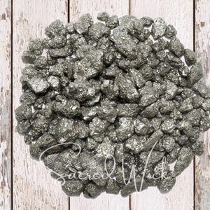 May include: A pile of raw pyrite stones, also known as fool's gold, with a metallic, silver-gray color. The stones are clustered together on a white wooden surface. The text "Sacred Wick" is visible in the lower portion of the image.