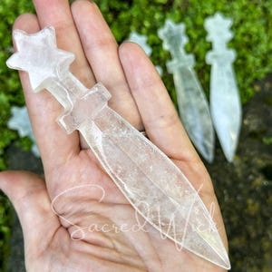 Clear quartz crystal athame resting on open palm, showing size with more athames on greenery in the background.