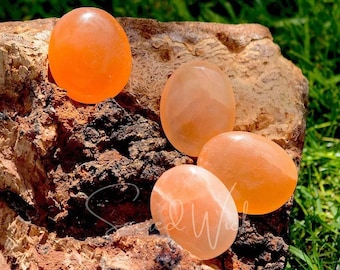 May include: Four smooth, polished orange carnelian stones, each with a slightly different shade of orange, are arranged on a rough, brown, textured surface.