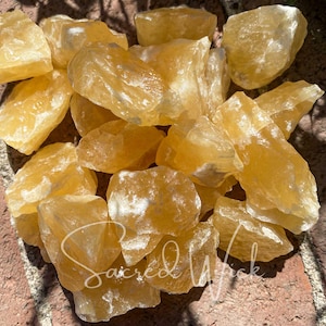 May include: A pile of rough, golden yellow calcite crystals. The crystals are translucent, with variations in colour and texture. The image is taken in natural light, highlighting the mineral's lustre. The text "Sacred Wick" is visible.