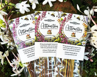 May include: Three packets of incense sticks, each labelled "Attraction" with floral and bee illustrations. The text reads "Wildflower Honey & Tahitian Vanilla". The background shows white flowers and green foliage, creating a natural aesthetic.