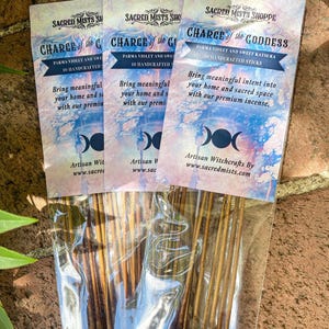 May include: Three packages of incense sticks with labels that read "Charge of the Goddess" and "Charge of the God". The labels feature a triple moon symbol and the text "Parma Violet and Sweet Katsura 10 Handcrafted Sticks". The incense sticks are packaged in clear plastic.