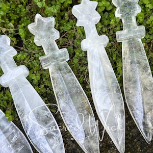 Close-up of six clear quartz crystal athames with star handles displayed on greenery in natural sunlight.
