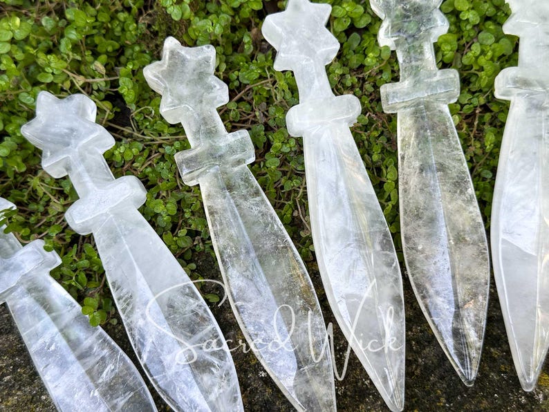 Close-up of six clear quartz crystal athames with star handles displayed on greenery in natural sunlight.