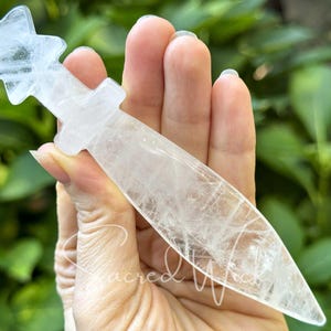 Clear quartz crystal athame with star handle held between fingers, greenery in the background.